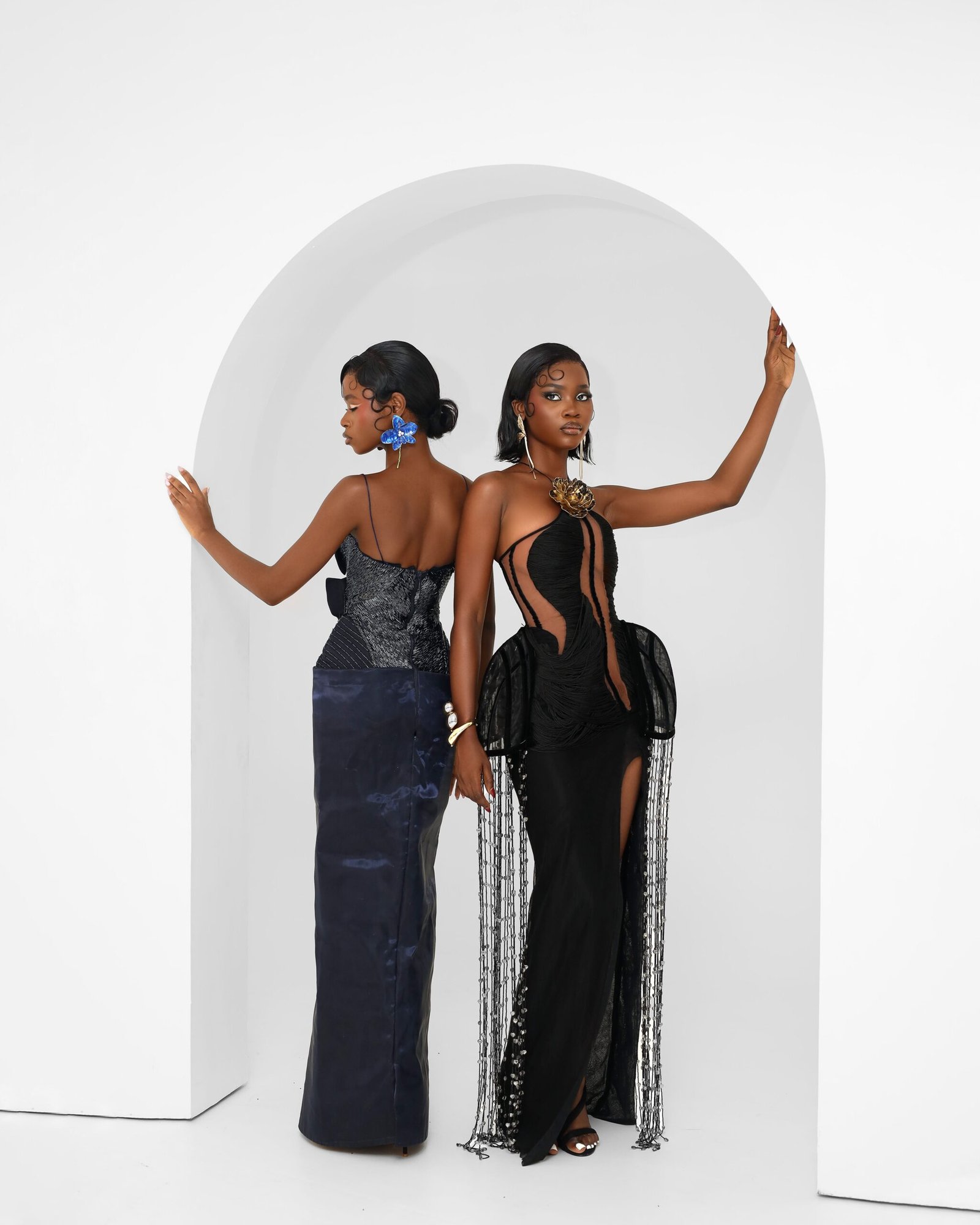 Two women in stunning black gowns posing elegantly in a minimalist studio archway.